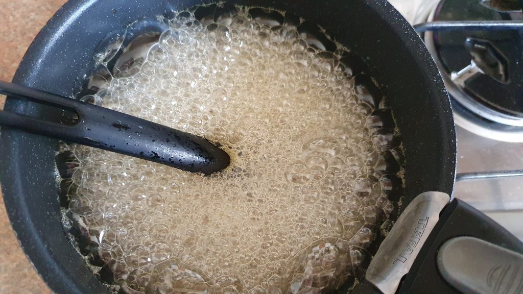lots of bubbles of sugary liquid
 in a pan with a dark plastic spoon handle