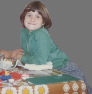 me as a 9 year old with a green shirt, smiling at the camera. i'm sitting at a table with some lego brick and a big green plaque
