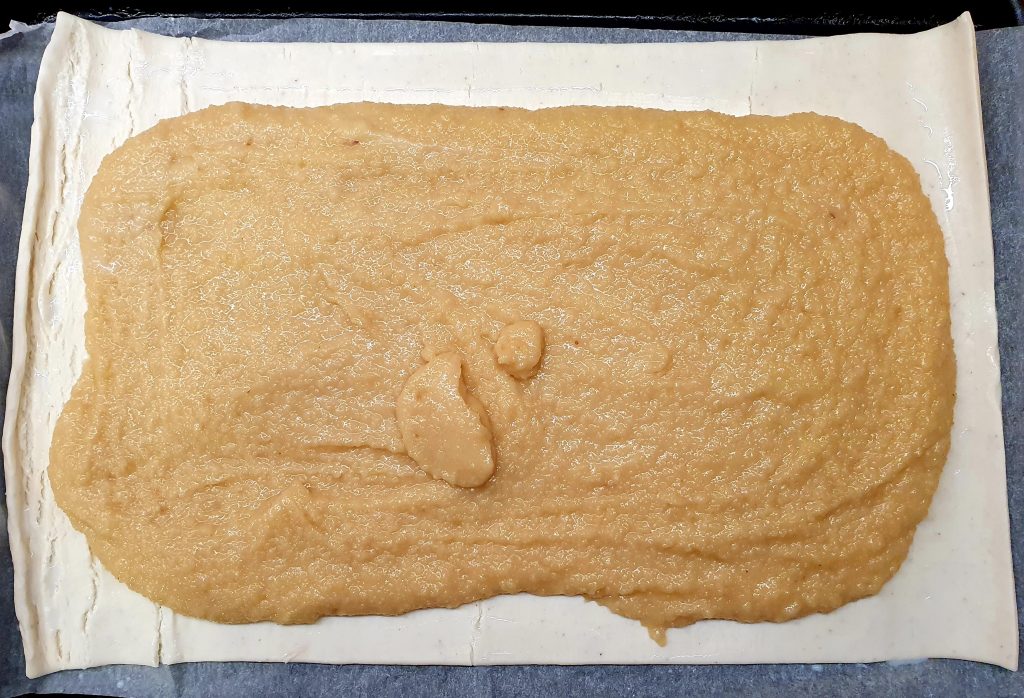 a light brown frangipane mix is spread on a rectangle of uncooked puff pastry. it stops about 2 cm from the edge and some of this part of the puff pastry is a bit wet with milk 