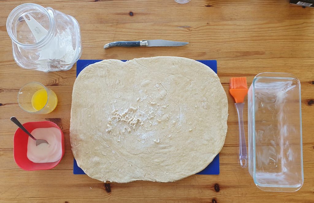 on a table the dough is spread in a rectangle, all that is needed is there around it: a glass with melted butter, a brush, a buttered cake tin, a bowl of sugar and a knife