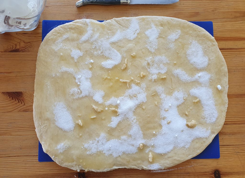 the rectangle of dough is shinier with the melted butter and the sugar is unevenly spread with some patch with more sugar