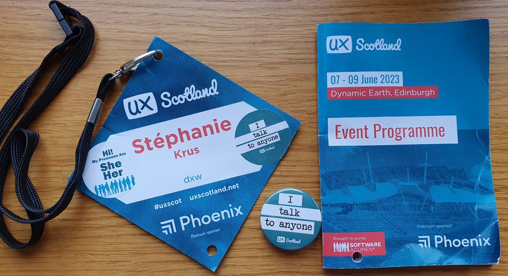 diamond shaped paper pass for the conference with a black lanyard, the little programme booklet and a badge saying" I talk to anyone'. On the pass there are logos, my name and where I work: dxw, a sticker with my pronouns she/her and another sticker saying that I talk to anyone