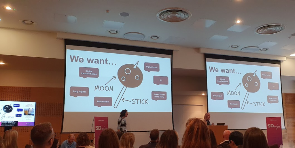the same slide displayed on 2 big screens and a small more colourful TV screen with two speakers in a big room. The slide shows a drawing for the moon on a stick to illustrate people wanting too many things