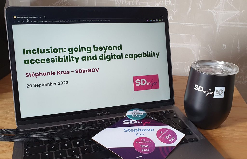 conference badge with my name and she/her stickers on the keyboard of an open laptop showing the first slide of my talk: Inclusion - going beyond accessibility and digital capability, there is a black cup next to it with the SDinGOV logo on it