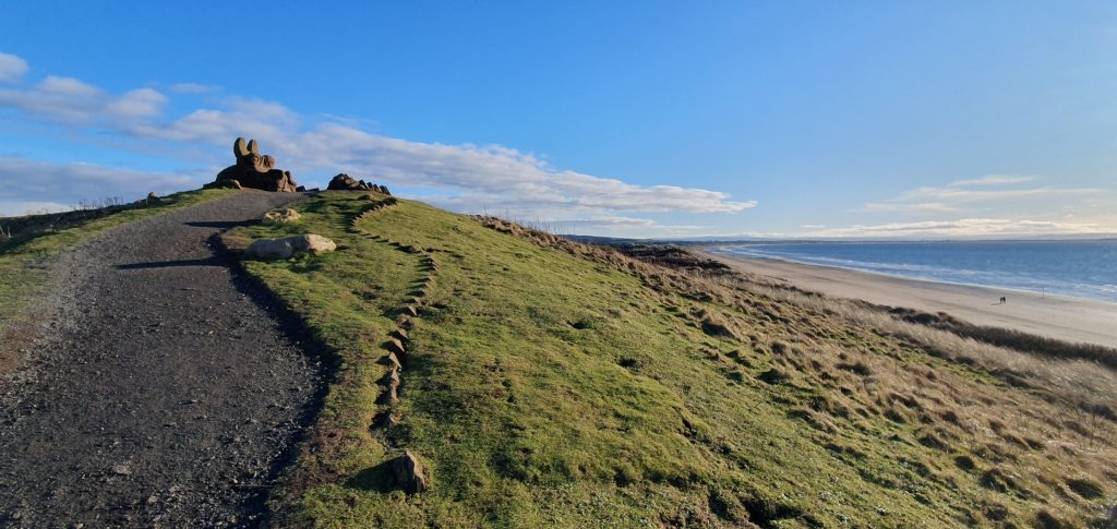 the head of a dragon made of rocks is seen at the top of a grassy dune  overseeing the beach below, the grey path up to the dragon's head has some pointy rocks part of its long wavy tail, it's a sunny day, the sky is mostly blue with some white and grey clouds