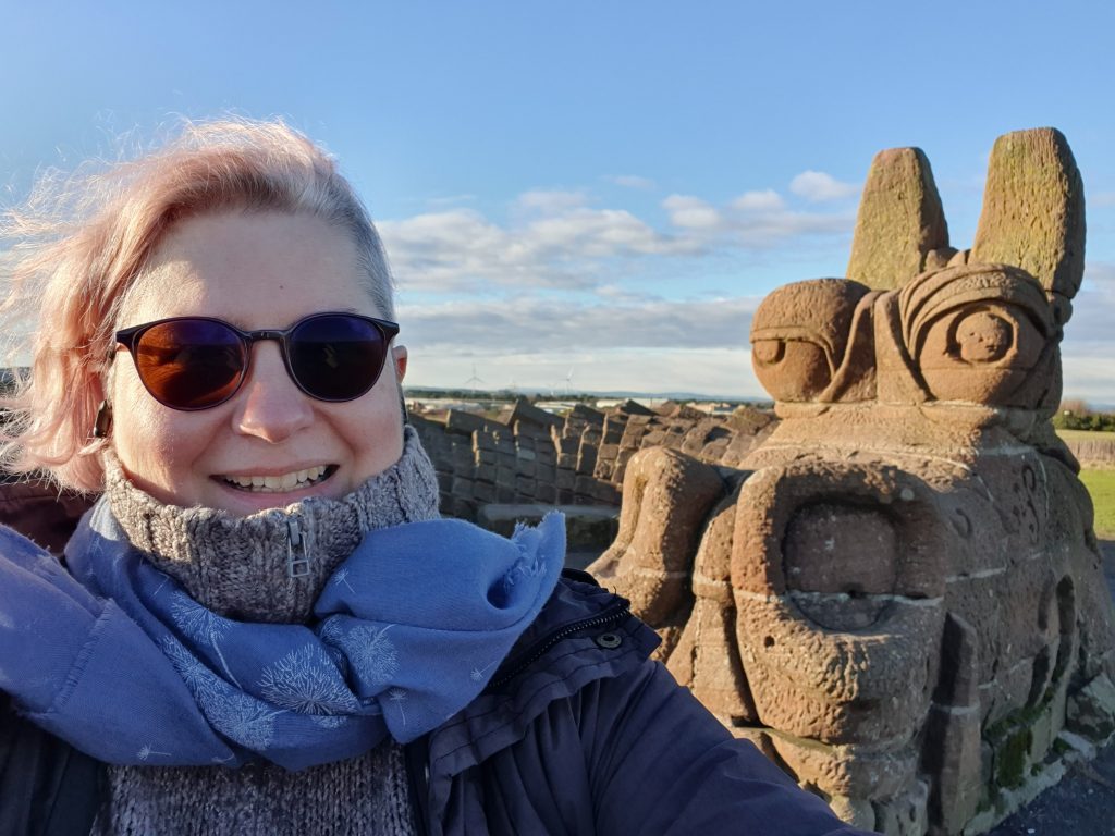 selfie with a close up of the rock dragon's head on the right and me on the left. I'm smiling, I have wind in my hair, my glasses are darken because of the sun, I have a wool top and a scarf which give you a feel that it's a sunny but cold day