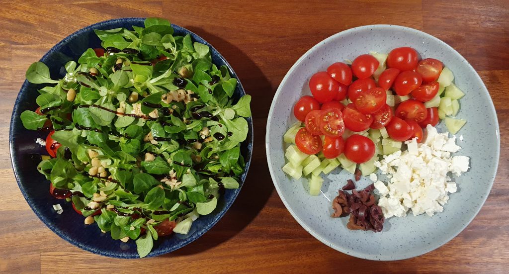 two plates , the left one has lamb lettuce and nuts on the top with lines of balsamic vinegar, hiding other veg underneath, the right one doesn't have the salad, so you can see the feta cheese, cherry tomatoes, cucumber and olives
