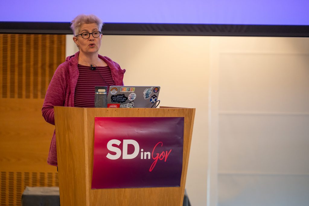I'm speaking, standing behind a pedestal with the SDinGOV logo, we can see my laptop on the pedestal with some stickers, and I'm wearing a mic on collar of my strippy teeshirt