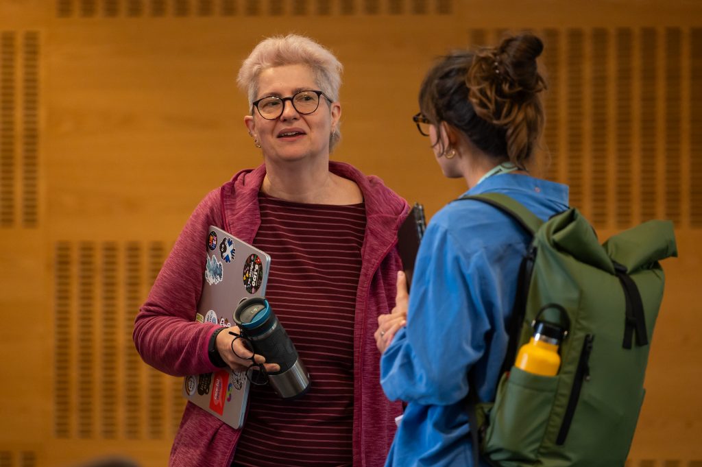 I'm standing and chatting with a person in the room after the talk. I'm facing, half smiling and holding my laptop, an extra pair of glasses and my water bottle. The other person has their back to the camera, is holding a black note book or laptop and has a bag pack with a water bottle