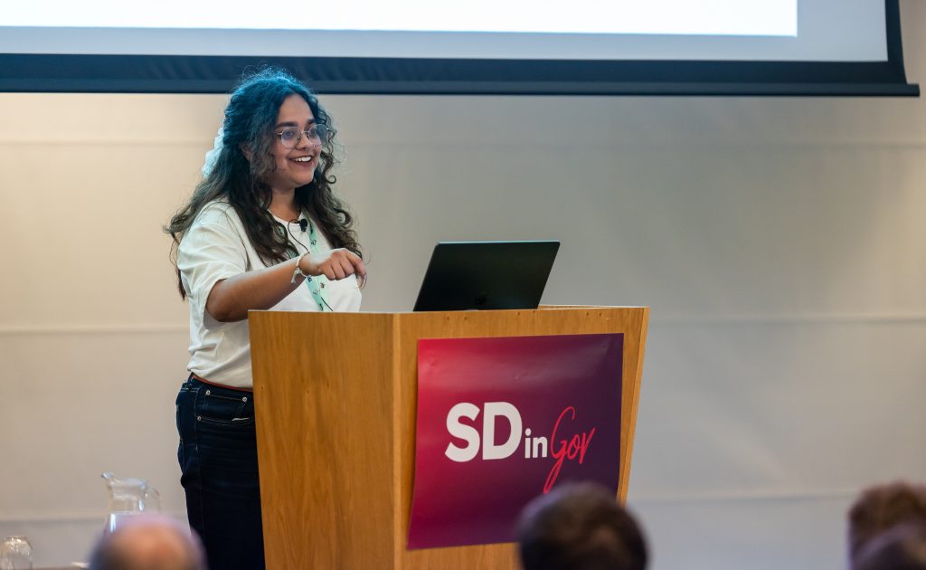 Bahana has a big smile and is standing on stage in front of a white screen and behind a wood pedestral with the SDinGOV logo. One of her hands is pointing to the audience. She is wearing  her conference lanyard over a white teeshirt and is wearing jeans and round glasses