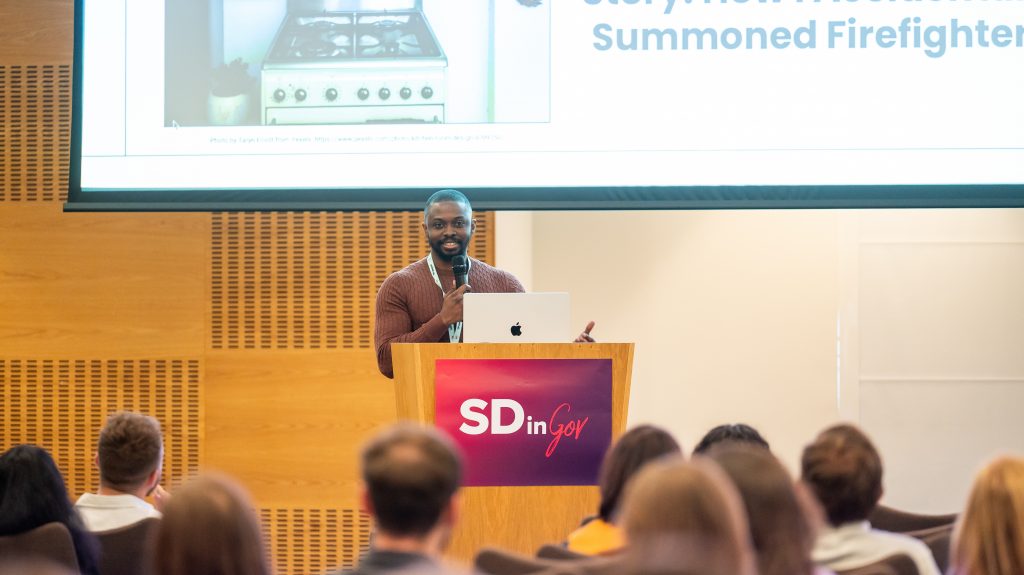 Kola is smiling while speaking on stage using a mic, standing behind a wood pedestal with the SDinGOV logo on it. We can see part of his laptop on it, and there is part of a slide displayed on a big screen behind him.