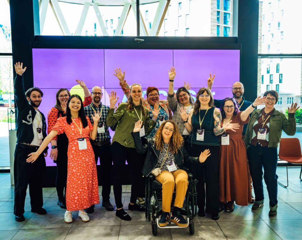 A group of eleven people pose together indoors in front of a large purple Access:Given display screen, smiling and waving at the camera. The group wear conference lanyards and colourful outfits, creating a warm, welcoming atmosphere in the bright modern venue with floor-to-ceiling windows.