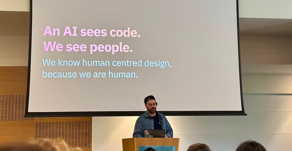 Craig on stage with a slide stating: An AI sees code. We see people. We know human centred design because we are human.
