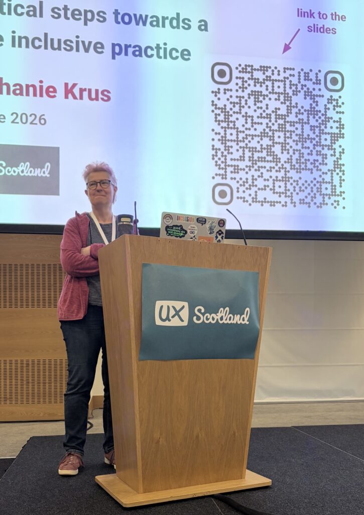 I'm on stage with my arms closed and a weird face, there is the pedestal in front of me with my laptop and a poster with with UX Scotland logo and at the back there is a slide with a QR code to get the link to my slides